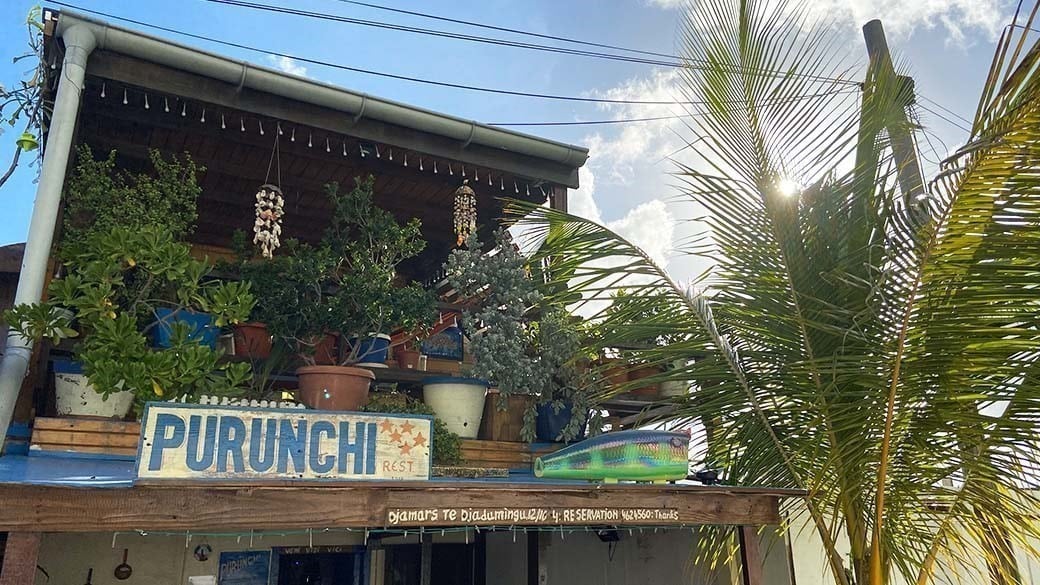 Der mit großen Topfpflanzen begrünte Balkon des Fischerhauses Purunchi - mit Windspielen, Namensschild, Öffnungszeiten und Telefonnummer